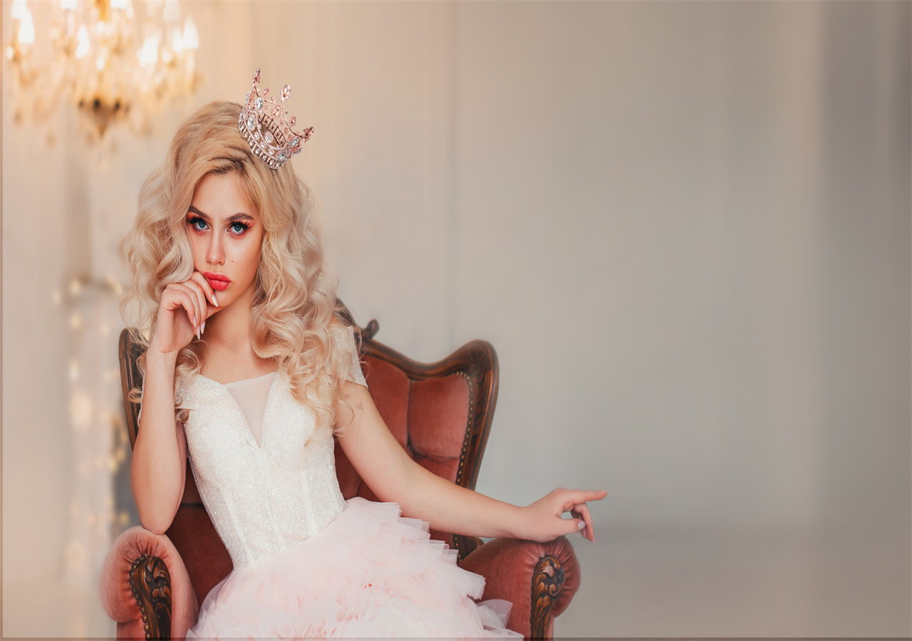 A girl with blond hair is wearing Sweetv crown. She sitting in vintage armchair. 